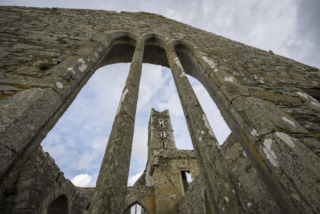 the looming remains of a grand window within the friary. Through the arches in the window where the glass would have been, you can see the tower of the friary in the near distance.