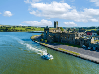 a bright blue day looking to the friary from an upward vantage point. A speedboat leaves ripples in the river passing by the friary.