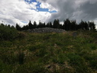 a stone cairn at the top of a grassy mound, backdropped by evergreen trees