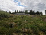 a stone cairn at the top of a grassy mound, backdropped by evergreen trees