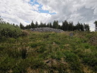 a stone cairn at the top of a grassy mound, backdropped by evergreen trees