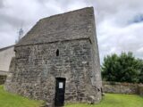 view of the entrance to the stone house. The entrance is a closed gate into a small rectangular style house, with a triangular roof.