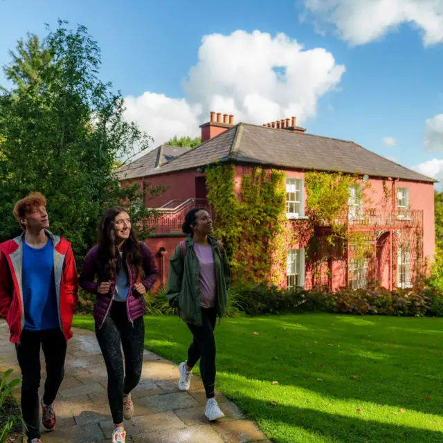 3 people visit a red coloured house with vines wrapped around its front exterior