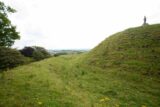 a raised mound in the grass, overlooking onto the distant landscape, with a statue standing on the top of the hill.