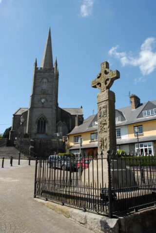 a high cross protected by a square fence in the middle of the town, and a high-steepled church on its left side.