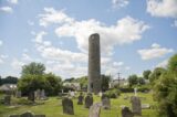 the round tower is almost touching the clouds floating in the sky, and stone graves surround the grounds it stands in.