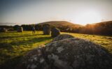 the sun sets behind the mountains in front of the stone circle. A man stands at the edge of the circle watching the sunset.