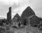 black and white image of the church ruins within a graveyard, with a partially disintegrating round tower to the left.