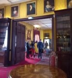 A tour Guide stands with others in the drawing room of Kilkenny Castle, the walls are yellow and hanged portraits line them.