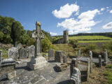 the high cross situated within the cemetery, surrounded by different size gravestones, with the church ruins in the background.