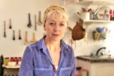A woman with short blonde hair poses for a photo in a blue shirt in a kitchen.