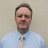 A man stands in a blue shirt and champagne coloured tie.
