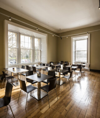 Internal image of Kilmainham Gaol Café showing 6 table and chairs with one large widow to the left and a smaller window just ahead of the seating.