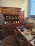 An old fashioned dresser with it's doors open with lots of jars and various kitchen ingredients on view on the dresser's shelves. In front of this dresser we are presented with a table covered in various kitchen utensils and bowls.