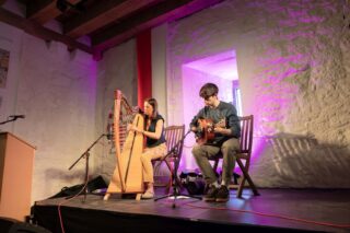 A lady sits and plays a harp along with a seatd man playing a guitar