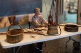 A lady sits at a table making a willow basket