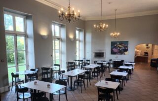 Tearoom with several tables and chairs windows to the left and an archway leading to a counter area