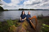 A harp is placed in front of a man and a lady who are sitting on the edge of a lake