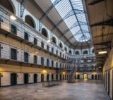 view of a spacious wing of Kilmainham Gaol in the evening, with the lights on, and the darkening sky visible through the glass ceiling above.
