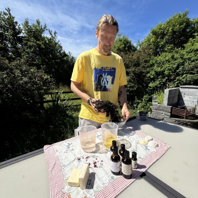 A man stands at a table holding seaweed. The table has ingredients for soap making
