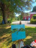 A painting sits on an easel in the outdoors with a tree and house in the background